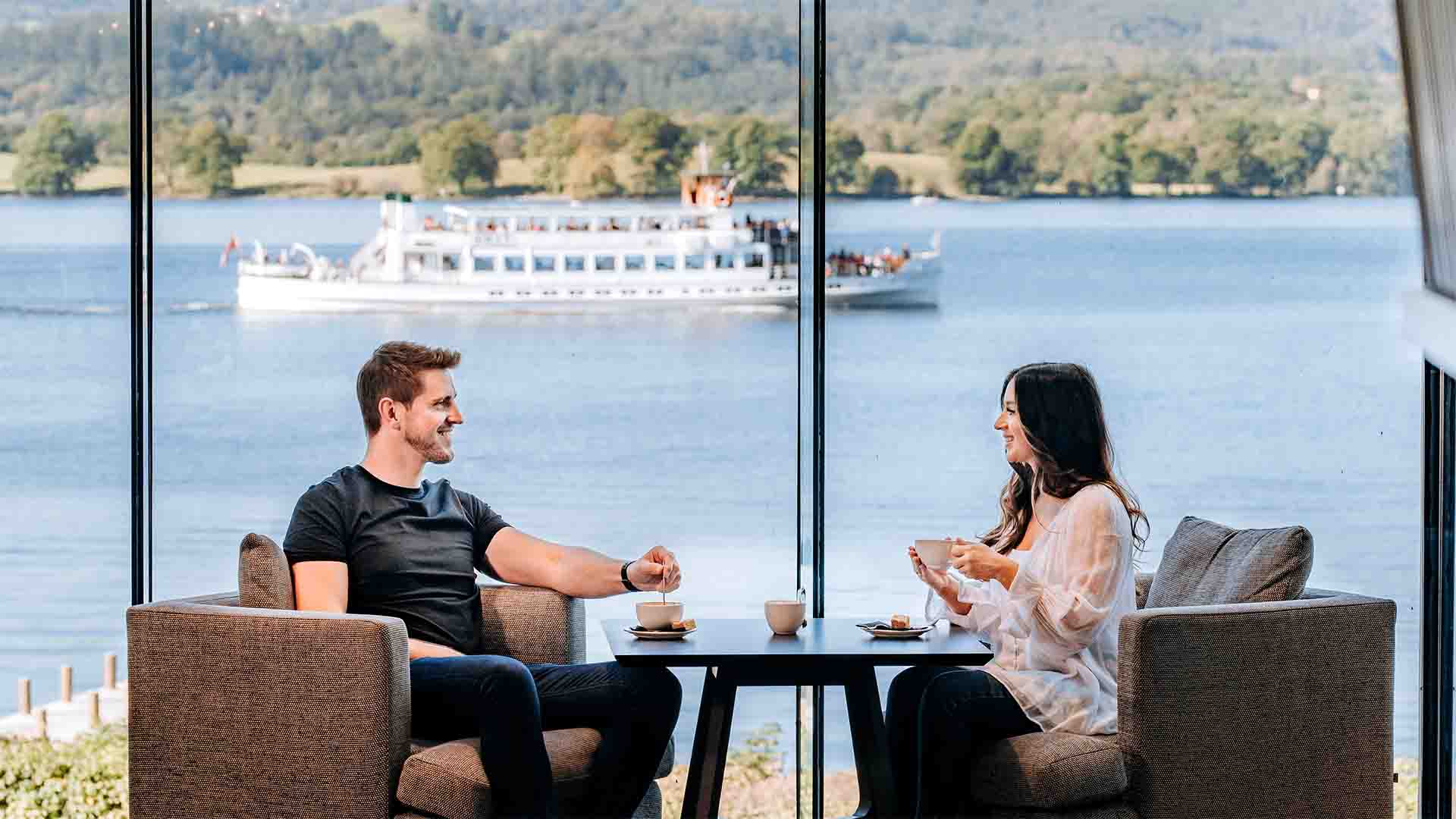 Couple sat in the hotel's Atrium with a Windermere lakes cruiser passing in the background 