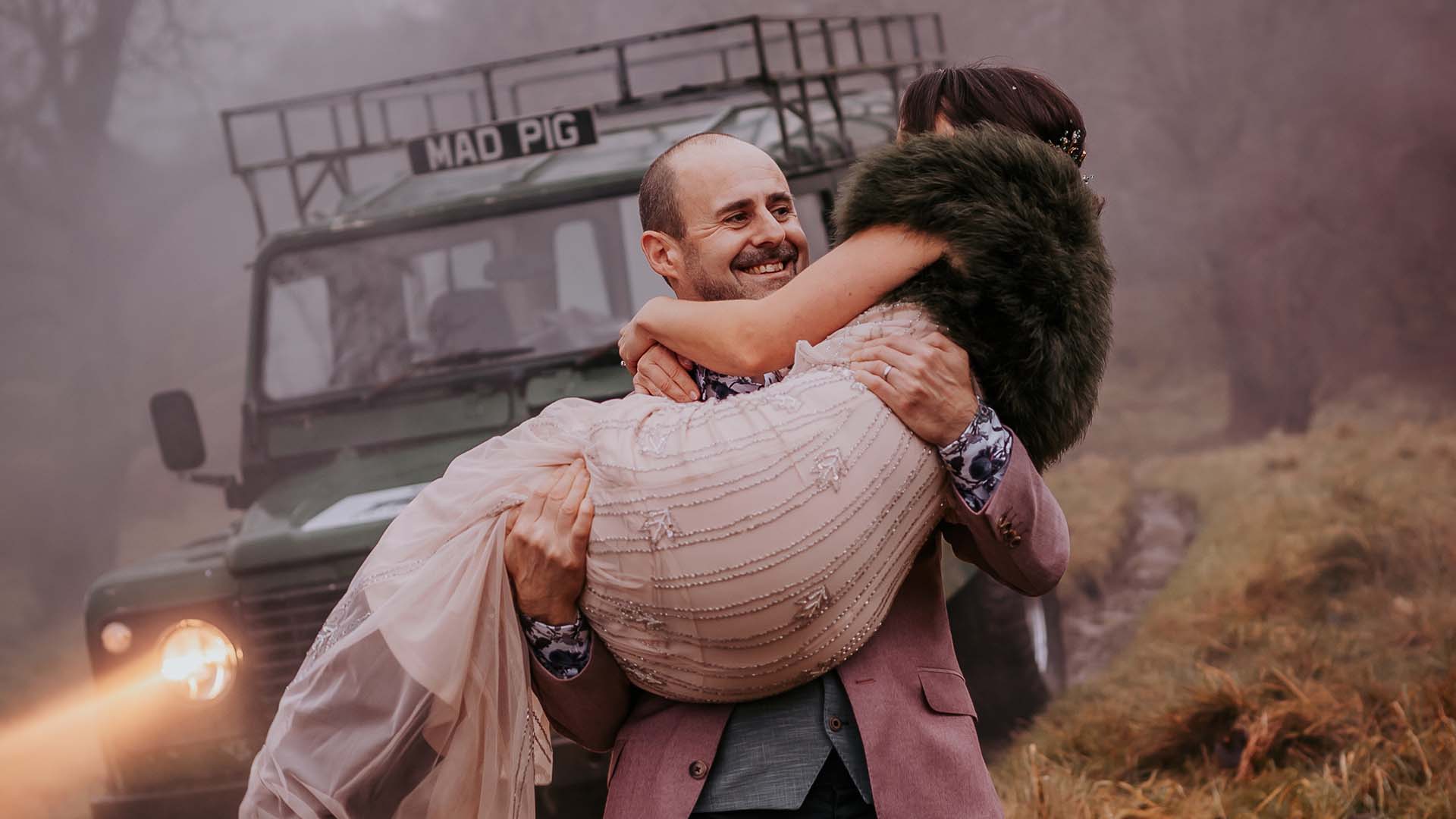 The groom carrying the bride with the Wild Boar Estate landrover behind