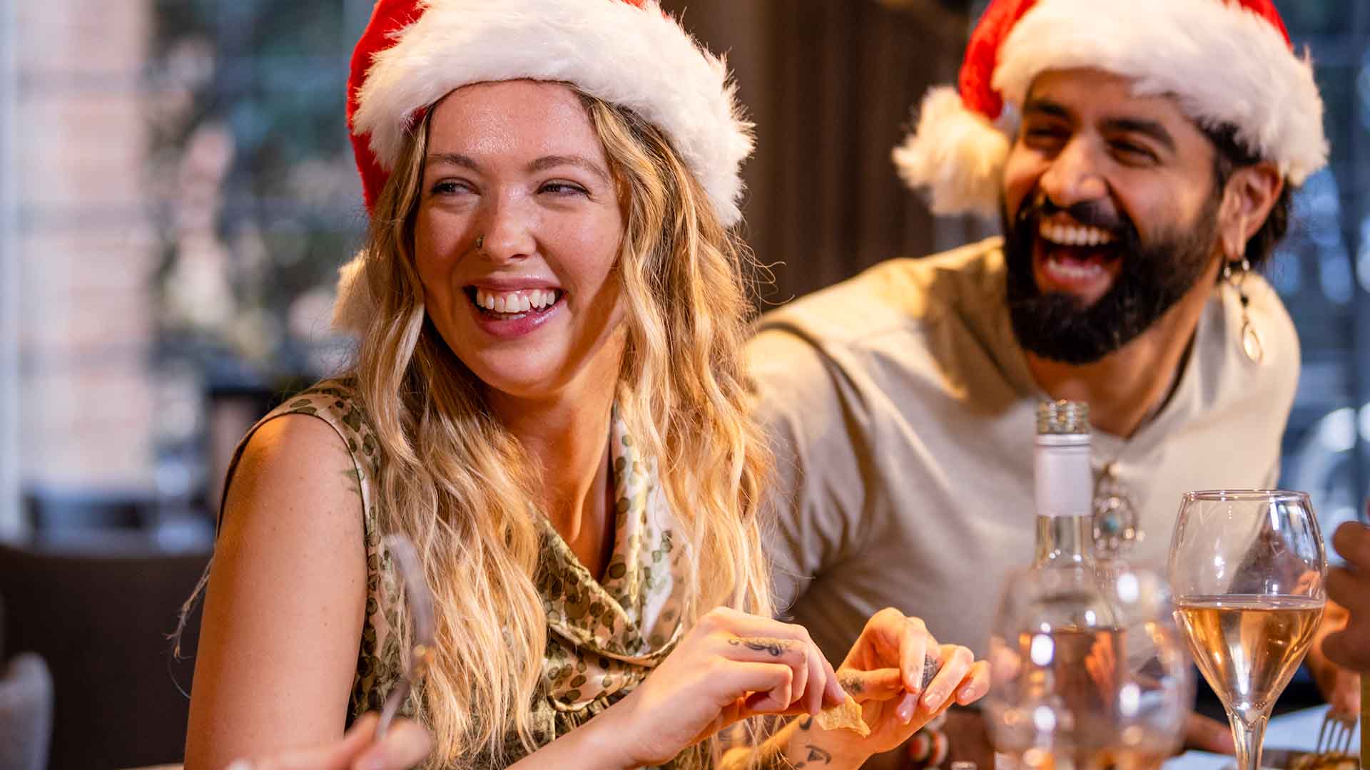 Two young adults wearing Christmas hats, laughing whilst seated at the table.