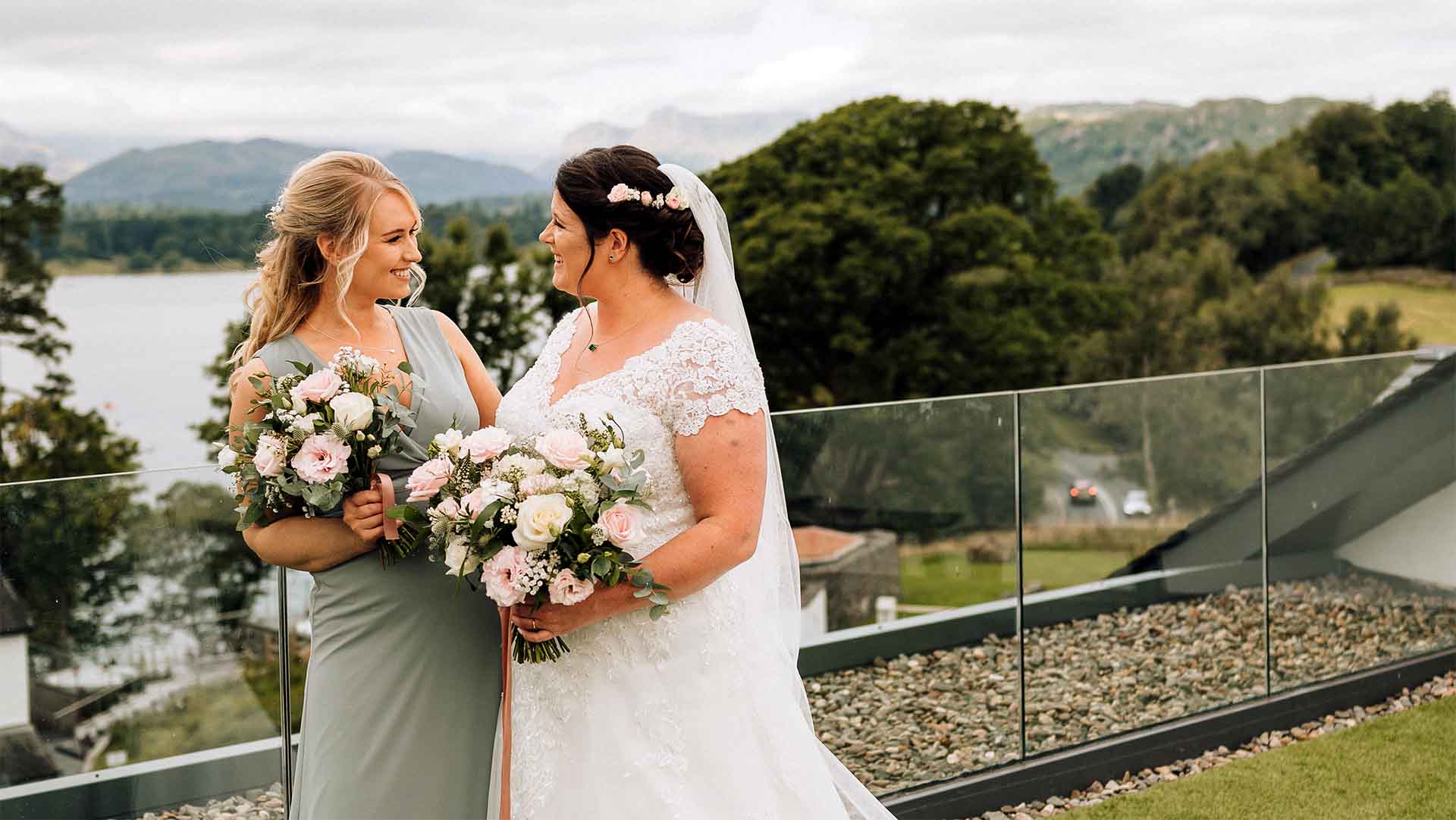 Bride and bridesmaid stood together on the Winander Rooftop Terrace with Windermere and Lake District fells in the background